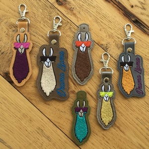 May include: Six colorful llama keychains with sunglasses and the text "Drama Llama" on a wood background.