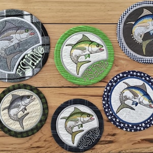 May include: Six round embroidered coasters featuring a fish design. The coasters have different colored backgrounds and borders. One coaster has the word "Fishing" embroidered on it. Another coaster has the name "Justin" embroidered on it.