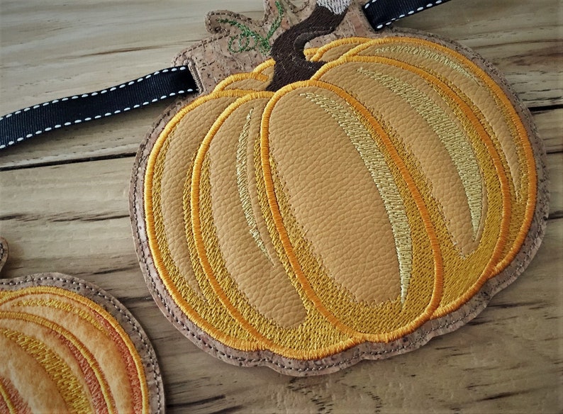 K&ouml;nnte beinhalten: Eine Herbstgirlande mit einem K&uuml;rbisdesign auf Korkunterlage. Der K&uuml;rbis ist mit gelbem und orangefarbenem Garn bestickt und hat einen braunen Stiel.