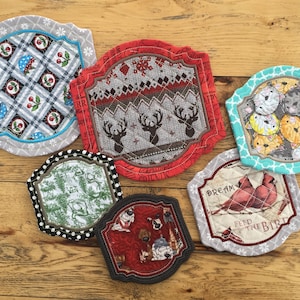 May include: Six different fabric pot holders with a variety of designs. The designs include a red and white checkered pattern with cherries, a red and white sweater pattern with reindeer, a teal pattern with cats, a green and white pattern with trees, a red pattern with dogs, and a gray pattern with cardinals and the text "Dream Feed the Bird".