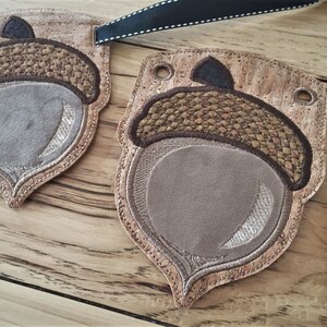 Puede incluir: Una pancarta en forma de bellota de color marr&oacute;n y beige con un respaldo de corcho. La pancarta est&aacute; decorada con detalles cosidos y tiene agujeros para colgar.