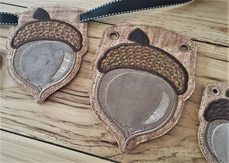 Puede incluir: Una pancarta en forma de bellota de color marr&oacute;n y beige con un respaldo de corcho. La pancarta est&aacute; decorada con detalles cosidos y tiene agujeros para colgar.