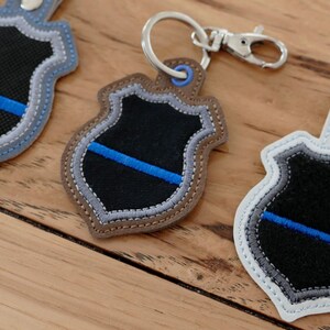 K&ouml;nnte beinhalten: Drei gestickte Schl&uuml;sselanh&auml;nger in Form eines Polizeibadges mit einem d&uuml;nnen blauen Streifen. Die Schl&uuml;sselanh&auml;nger sind aus schwarzem, grauem und wei&szlig;em Stoff gefertigt und haben einen silbernen Metallring.