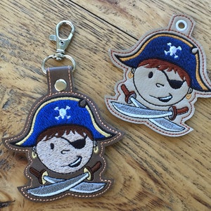 Puede incluir: Dos llaveros de cuero marrón con imágenes bordadas de un pirata de dibujos animados con un sombrero azul, un parche en el ojo y espadas cruzadas. El pirata tiene una calavera y huesos cruzados en su sombrero.