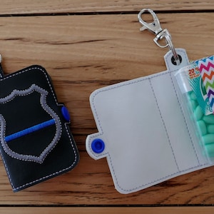 May include: Two keychains with different designs. The keychain on the left has a black leather case with a gray and blue embroidered police badge. The keychain on the right has a white leather case with a clear plastic pocket for holding a small container of mints. The mints are green and wrapped in a colorful chevron pattern.