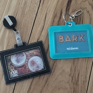 Puede incluir: Dos porta-credenciales sobre una superficie de madera. Uno es negro con una foto de cerveza y el texto "Beer Store". El otro es turquesa con la palabra "BARK" y el logo de PetSmart. Ambos tienen ventanas transparentes.