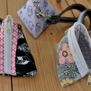 May include: Three triangular coin purses with zippers and wrist straps. One is black with white feather patterns, another is floral, and the third is light purple with a pattern. The purses are open, revealing the interior fabric.