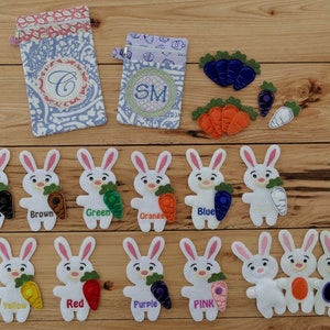 Op de afbeelding: Een set van vingerpoppen van vilt in de vorm van konijnen met wortels in verschillende kleuren, waaronder zwart, bruin, groen, oranje, blauw, geel, rood, paars, roze en wit. De poppen zijn in twee rijen op een houten oppervlak gerangschikt. Er zijn ook twee kleine zakjes met de geborduurde letters 'C' en 'SM'.