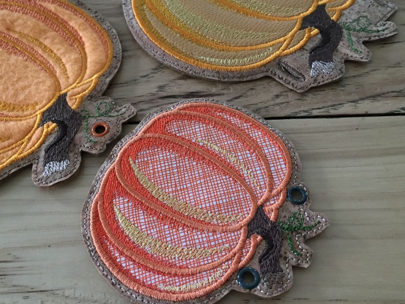 K&ouml;nnte beinhalten: Drei gestickte Stoff-Ornamente in Form von K&uuml;rbissen mit braunen Stielen und gr&uuml;nen Bl&auml;ttern. Die K&uuml;rbisse sind orange, gelb und rot mit einem strukturierten Design.