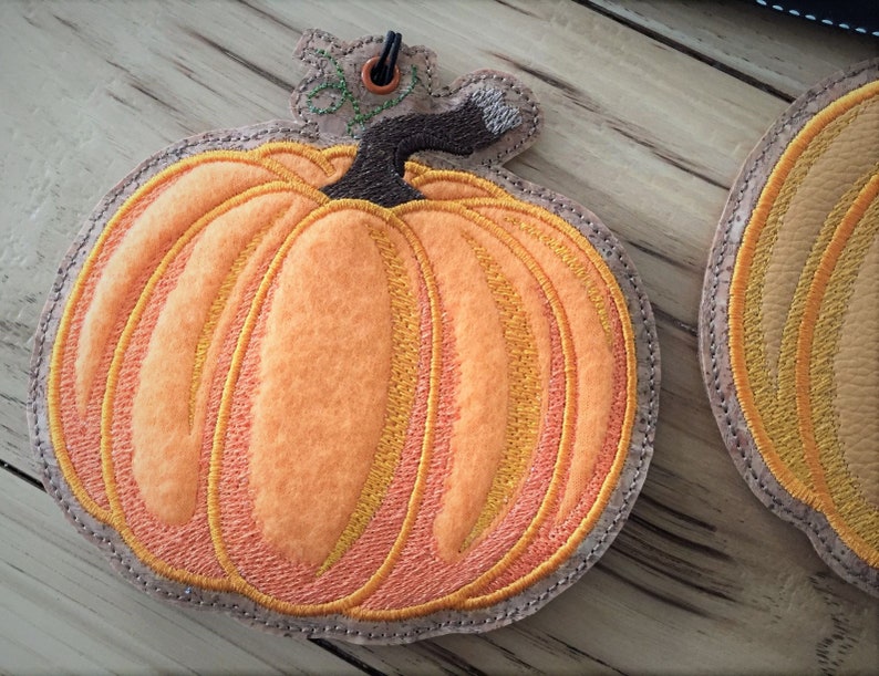 K&ouml;nnte beinhalten: Orangefarbener Filz-K&uuml;rbis-Anh&auml;nger mit braunem Stiel und gr&uuml;nen Bl&auml;ttern. Der K&uuml;rbis hat ein gesticktes Design mit orangem und gelbem Garn.