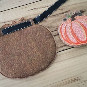 K&ouml;nnte beinhalten: Zwei Filz-K&uuml;rbisse, einer orange und einer braun, mit einem schwarzen Band, das an dem braunen K&uuml;rbis befestigt ist. Der orange K&uuml;rbis hat einen gr&uuml;nen Stiel und ist mit einem detaillierten Muster bestickt.