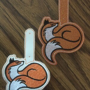K&ouml;nnte beinhalten: Zwei bestickte Gep&auml;ckanh&auml;nger in Fuchsform. Ein Anh&auml;nger ist orange mit wei&szlig;em Bauch und wei&szlig;em Rand. Der andere Anh&auml;nger ist orange mit Glitzer-Finish und wei&szlig;em Rand. Beide haben einen wei&szlig;en Riemen.