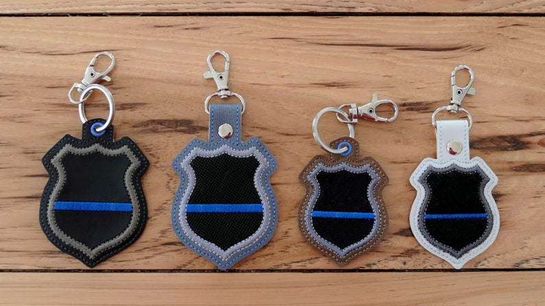 K&ouml;nnte beinhalten: Vier Schl&uuml;sselanh&auml;nger mit einem blauen Linien-Polizeibadge-Design. Die Schl&uuml;sselanh&auml;nger sind aus Leder gefertigt und haben einen silbernen Metallring. Die Abzeichen sind in verschiedenen Farben: Schwarz, Grau, Braun und Wei&szlig;.