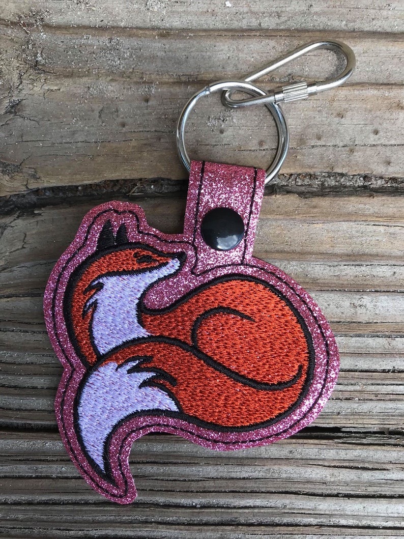 K&ouml;nnte beinhalten: Ein pinkfarbener Glitzer-Fuchs-Schl&uuml;sselanh&auml;nger mit einem silbernen Schl&uuml;sselring. Der Fuchs ist mit orangefarbenem, schwarzem und lavendelfarbenem Faden bestickt. Der Schl&uuml;sselanh&auml;nger hat einen schwarzen Druckknopfverschluss.