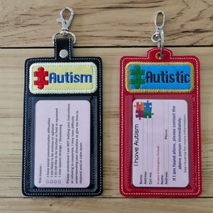May include: Two black and red leather ID card holders with clear plastic windows. The top of each holder has a blue and yellow puzzle piece with the words "Autism" and "Autistic" embroidered on them. The card holders have a metal ring for attaching to a keychain.