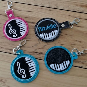 May include: Four round keychains with black and white piano keys and a treble clef design. One keychain is pink, one is black, one is blue, and one is teal. The black keychain has the name "Amélie" embroidered on it.