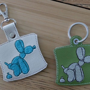 May include: Two keychains with a balloon animal design. One keychain is white with a blue balloon animal and the other is green with a gray balloon animal.