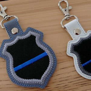 K&ouml;nnte beinhalten: Zwei Schl&uuml;sselanh&auml;nger mit einer blauen Linie auf einem schwarzen Schild. Ein Schl&uuml;sselanh&auml;nger ist grau und der andere ist wei&szlig;.