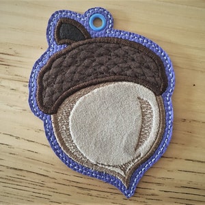 Puede incluir: Un adorno de bellota de fieltro marr&oacute;n y blanco con un lazo azul para colgar. La bellota tiene una tapa marr&oacute;n y un cuerpo blanco con costuras marrones.