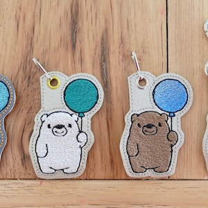 Peut inclure: Quatre porte-clés brodés représentant un ours tenant un ballon. Les ours sont marron, blanc, marron et gris. Les ballons sont bleus, verts, bleus et roses.