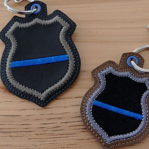K&ouml;nnte beinhalten: Zwei schwarze Leder-Schl&uuml;sselanh&auml;nger in Form von Polizeisternchen mit einem blauen Streifen in der Mitte. Die Schl&uuml;sselanh&auml;nger haben einen silbernen Metallring und einen silbernen Metallverschluss.