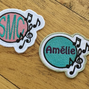 Puede incluir: Dos parches de planchado bordados con notas musicales. Un parche es rosa con las iniciales "SMC" en turquesa. El otro parche es turquesa con el nombre "Amélie" en morado.