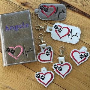 Puede incluir: Un conjunto de seis llaveros con un corazón rojo con una huella de pata negra y una línea de latido del corazón. Los llaveros están hechos de cuero blanco y gris. Un llavero está unido a una libreta de cuero gris con el nombre "Angela" bordado en la parte delantera.