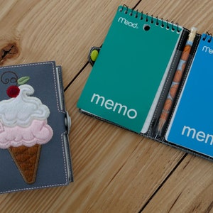 Puede incluir: Una cubierta de cuaderno de tela gris con un diseño de cono de helado bordado. La cubierta tiene un bolsillo en la parte delantera que contiene dos pequeños cuadernos de espiral. Los cuadernos son azul y verde y tienen la palabra "memo" impresa en ellos.
