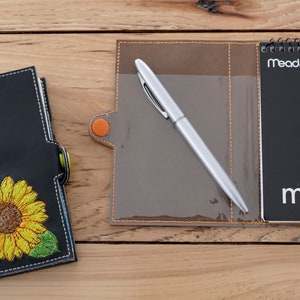 Op de afbeelding: Een zwart lederen notitieboek met een geborduurd zonnebloemontwerp en een bijpassende bruine leren pennenhouder met een zilveren pen. De pennenhouder heeft een klein notitieblok met de tekst "Mead. memo" op de voorkant.