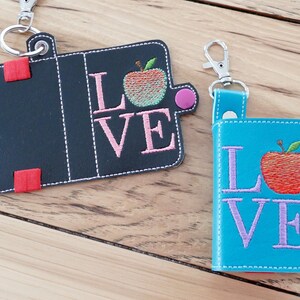 Puede incluir: Dos llaveros con cubiertas de imitación de cuero negro y azul. Ambos llaveros tienen una manzana roja y la palabra "LOVE" bordada. La manzana del llavero negro es verde y rosa, mientras que la manzana del llavero azul es roja.