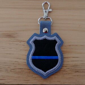 K&ouml;nnte beinhalten: Ein blauer und silberner gestickter Schl&uuml;sselanh&auml;nger in Form eines Polizeibadges mit einem d&uuml;nnen blauen Streifen in der Mitte.
