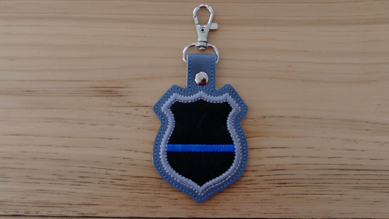 K&ouml;nnte beinhalten: Ein blauer und silberner gestickter Schl&uuml;sselanh&auml;nger in Form eines Polizeibadges mit einem d&uuml;nnen blauen Streifen in der Mitte.