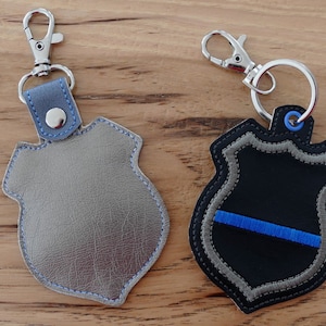 K&ouml;nnte beinhalten: Zwei Leder-Schl&uuml;sselanh&auml;nger in Form von Polizeiwappen. Einer ist grau mit blauen N&auml;hten und der andere ist schwarz mit einem blauen Streifen und grauen N&auml;hten.