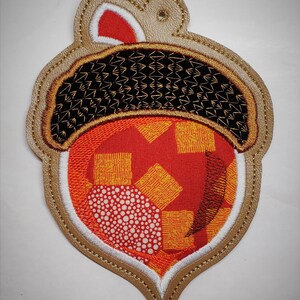 Puede incluir: Un adorno de fieltro marr&oacute;n en forma de bellota con un centro de tela con estampado rojo y naranja. La bellota tiene una tapa de fieltro blanco y un dise&ntilde;o de hilo dorado.