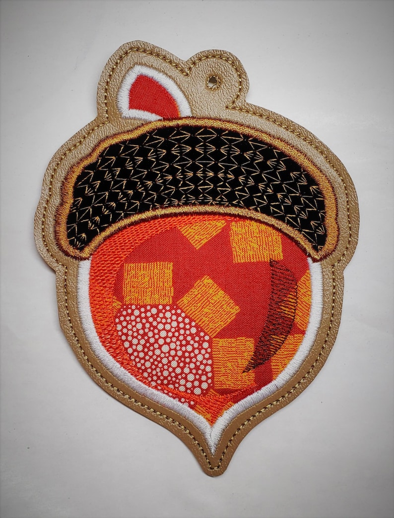 Puede incluir: Un adorno de fieltro marr&oacute;n en forma de bellota con un centro de tela con estampado rojo y naranja. La bellota tiene una tapa de fieltro blanco y un dise&ntilde;o de hilo dorado.