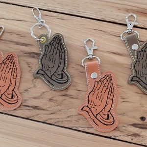 May include: Four leather keychains with a design of praying hands. The keychains are in brown, tan, and gray.