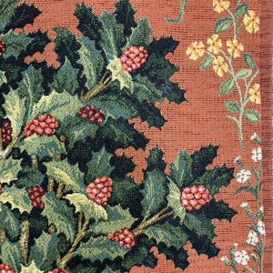 Op de afbeelding: Close-up van een wandtapijt met een gedetailleerd ontwerp van een groene, bladerrijke plant met rode bessen tegen een warme oranje achtergrond. De plant is geweven met ingewikkelde details, waaronder donkergroene bladeren en kleine rode bessen.