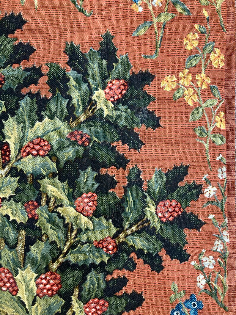 Op de afbeelding: Close-up van een wandtapijt met een gedetailleerd ontwerp van een groene, bladerrijke plant met rode bessen tegen een warme oranje achtergrond. De plant is geweven met ingewikkelde details, waaronder donkergroene bladeren en kleine rode bessen.