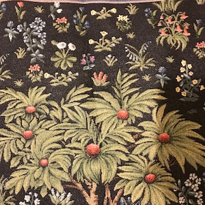 Op de afbeelding: Een wandkleed met een gestileerde boom met groene bladeren en rode vruchten. De boom is omringd door een verscheidenheid aan bloemen en planten in tinten blauw, geel, roze en wit. De achtergrond is donkerbruin.