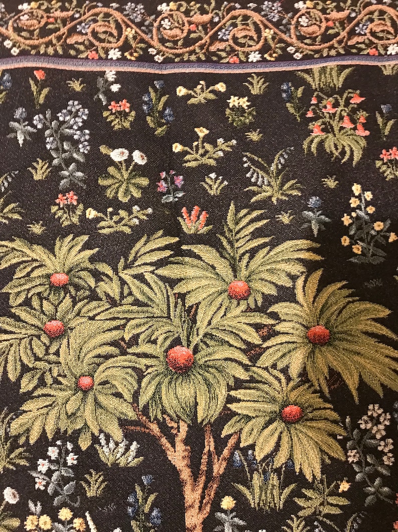 Op de afbeelding: Een wandkleed met een gestileerde boom met groene bladeren en rode vruchten. De boom is omringd door een verscheidenheid aan bloemen en planten in tinten blauw, geel, roze en wit. De achtergrond is donkerbruin.