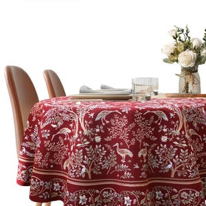 Könnte beinhalten: Eine runde, weinrote Tischdecke mit einem detaillierten weißen und goldenen botanischen Muster mit Vögeln und Bäumen. Die Tischdecke ist über einem Tisch drapiert, auf dem zwei braune Stühle zu sehen sind. Eine Vase mit weißen Rosen steht auf dem Tisch.