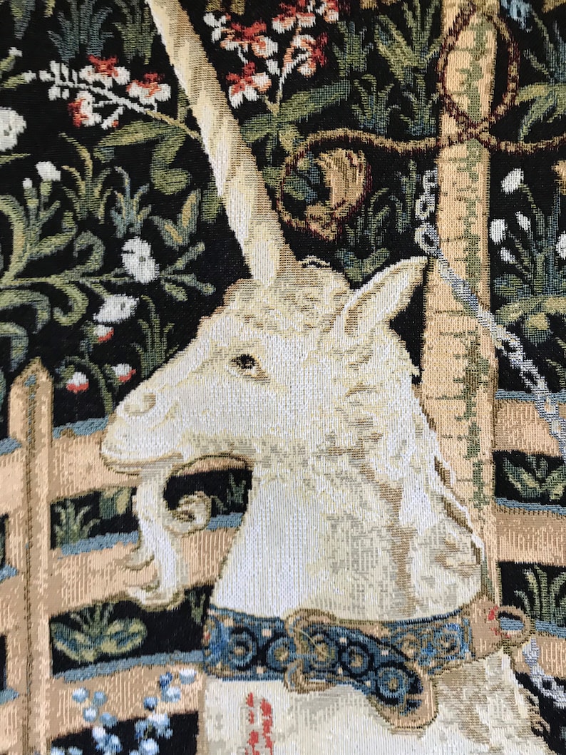 Puede incluir: Primer plano de un tapiz de unicornio blanco con un collar azul y dorado. El unicornio est&aacute; de pie en un entorno de bosque con una valla de madera y flores.
