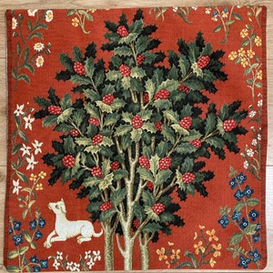 Op de afbeelding: Een wandkleed met een gestileerde boom met rode bessen en groene bladeren, omringd door verschillende bloemen. Een witte hond ligt in de rechterbenedenhoek van het wandkleed.