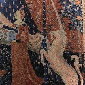 May include: A tapestry depicting a unicorn with a long, white horn, standing on its hind legs and resting its front legs on a tree trunk. The unicorn is white with a pink hue and is surrounded by a dark blue and red background with floral patterns.