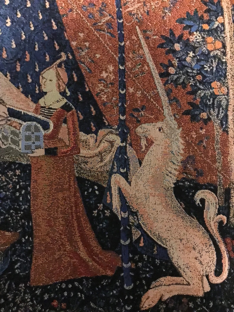 May include: A tapestry depicting a unicorn with a long, white horn, standing on its hind legs and resting its front legs on a tree trunk. The unicorn is white with a pink hue and is surrounded by a dark blue and red background with floral patterns.