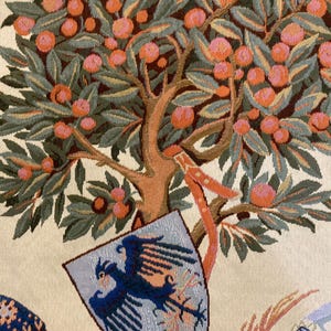 May include: A tapestry featuring a stylized tree with orange fruit and green leaves. A shield with a blue eagle is hanging from the tree. The background is a cream color.