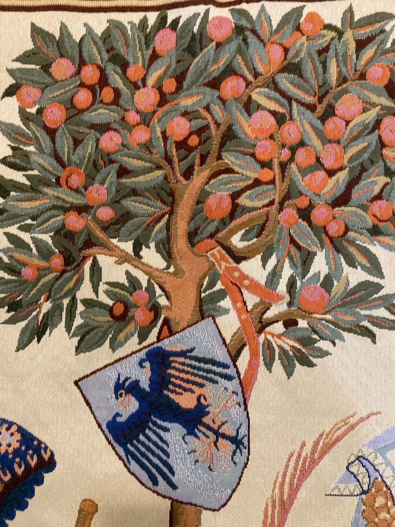 May include: A tapestry featuring a stylized tree with orange fruit and green leaves. A shield with a blue eagle is hanging from the tree. The background is a cream color.