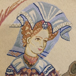 May include: A tapestry featuring a woman with a blue and white headdress, wearing a colorful, patterned dress. The tapestry has a cream background and is woven with intricate details.