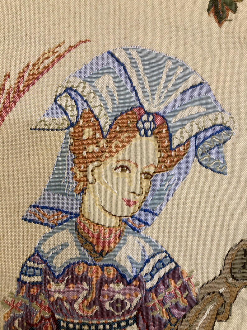 May include: A tapestry featuring a woman with a blue and white headdress, wearing a colorful, patterned dress. The tapestry has a cream background and is woven with intricate details.