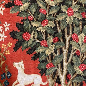 Op de afbeelding: Een wandkleed met een witte hond die ligt voor een rode achtergrond met een groene boom met rode bessen. De boom bevindt zich in het midden van de afbeelding en de hond in de linkerbenedenhoek.